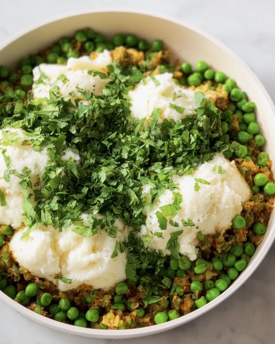 The image shows a close-up of a white pan filled with a layered dish. The bottom layer consists of finely cooked, golden-brown bits mixed with green herbs. On top of this layer, there are fresh bright green peas spread evenly. Large, soft white dollops of mashed potatoes sit on the peas, creating a fluffy texture contrast. Fresh green chopped cilantro is sprinkled around and mixed with the peas. The pan is set against a white marbled surface, and overall the colors are warm and fresh, showing a mix of green, white, and brown tones. photo taken with an iphone --ar 4:5 --v 7