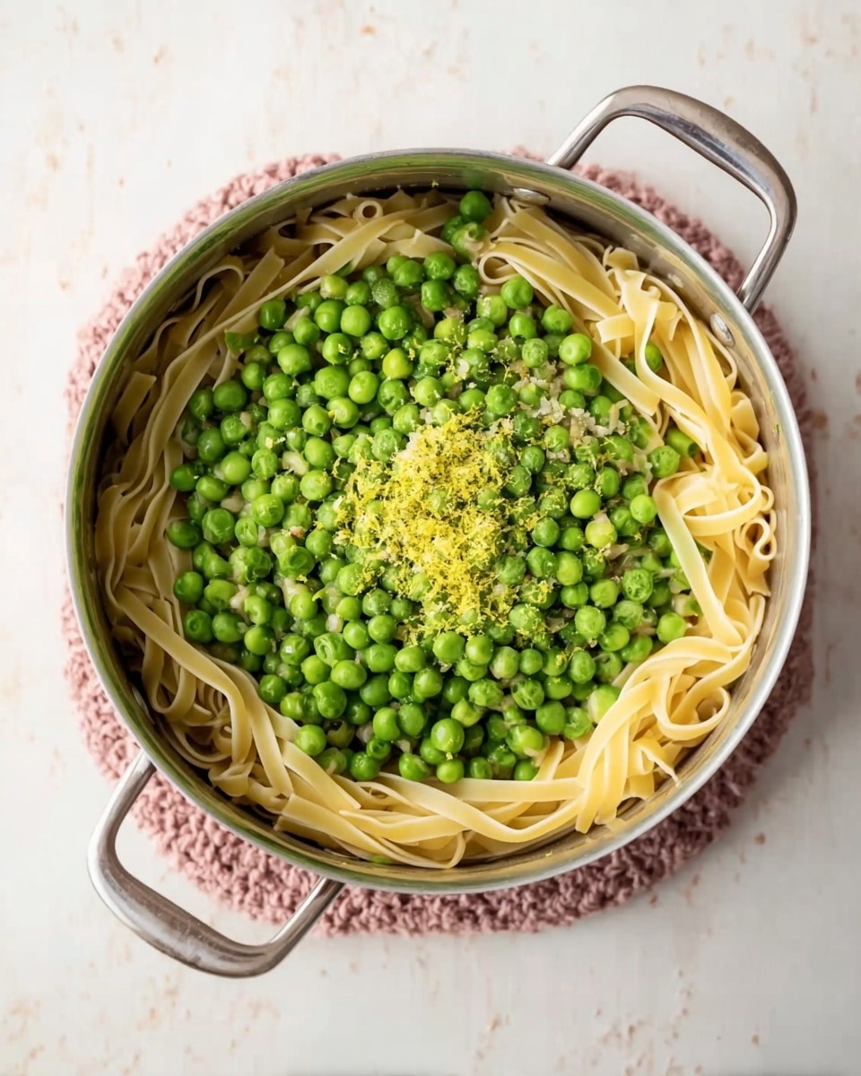 A metal pot is filled with two visible layers. The bottom layer consists of wide, pale yellow pasta strips placed loosely in a circular shape along the edge of the pot. On top of the pasta is a thick layer of bright green peas covering almost the entire surface. In the center of the peas, there is a small sprinkle of light yellow zest and a few bits of coarse salt. The pot sits on a soft knitted pink trivet on a white marbled texture surface. Photo taken with an iphone --ar 4:5 --v 7
