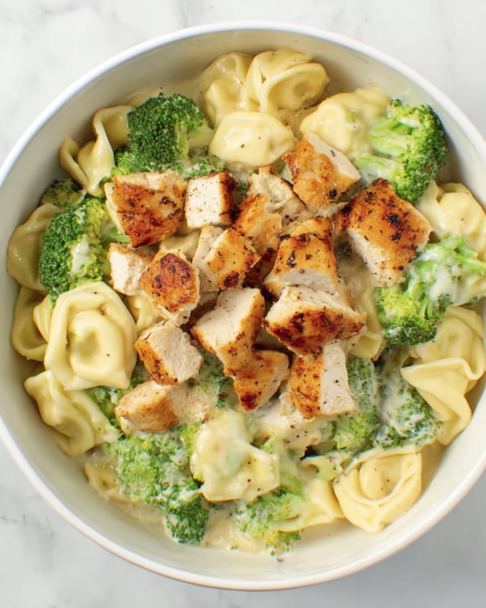 The dish shows a white bowl filled with three main layers. The bottom layer is a creamy, light yellow tortellini pasta, soft and smooth in texture. Mixed throughout this layer are bright green broccoli florets, fresh and slightly textured. On the top is a generous amount of chopped grilled chicken, golden brown with a slightly crispy outer surface and tender inside, spread evenly across the pasta and broccoli. The bowl sits on a white marbled surface. photo taken with an iphone --ar 4:5 --v 7