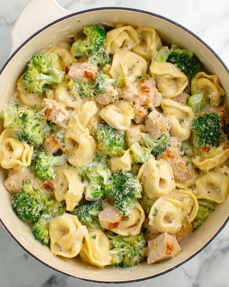 The image shows a white pot filled with a creamy pasta dish. The dish has three main layers: light yellow tortellini pasta, bright green broccoli florets, and chunks of light brown cooked chicken, all mixed evenly throughout. The creamy sauce coats every piece, giving a smooth texture to the whole dish. The background surface is white with a marbled texture. Photo taken with an iphone --ar 4:5 --v 7