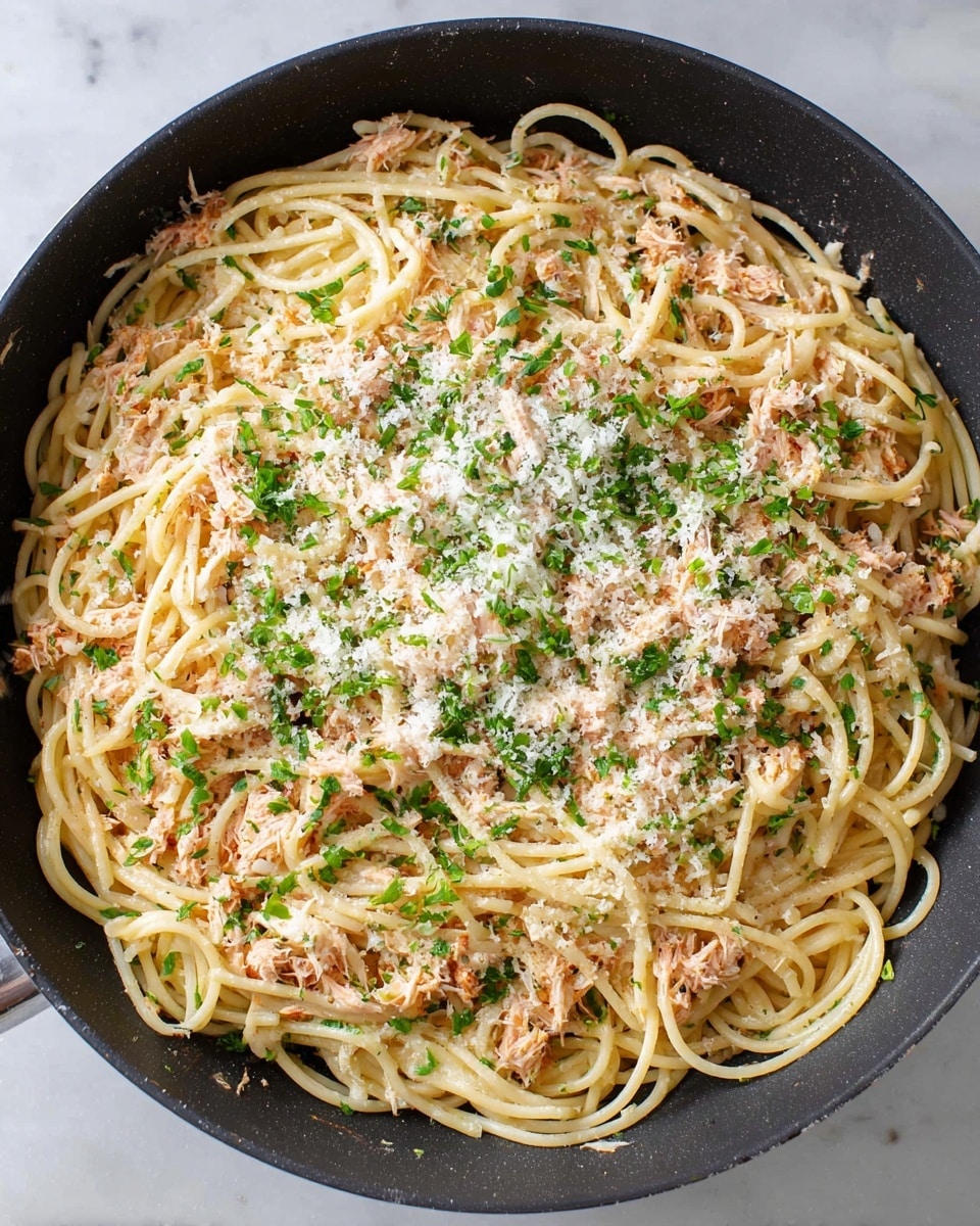 A close-up view of a black pan filled with a single layer of spaghetti pasta, mixed evenly with shredded light pink tuna. The top layer is sprinkled with finely grated white cheese and small chopped green herbs, mostly covering the pasta and tuna below. The pan sits on a white marbled surface, showing a simple, fresh pasta dish ready to serve photo taken with an iphone --ar 4:5 --v 7