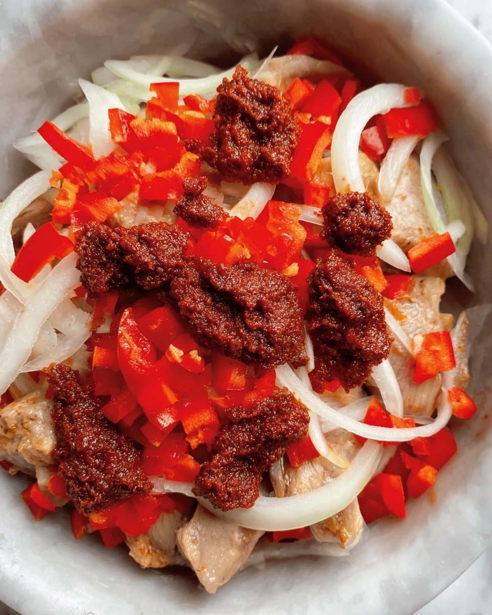 A close-up view of a dish in a white bowl showing three layers: the bottom layer is light beige pieces of meat, covered by a middle layer of thin white onion slices spread evenly, and topped with bright red chopped bell peppers scattered across the surface. On top, there are several dollops of dark reddish-brown thick paste, placed irregularly over the vegetables. The textures range from smooth and soft meat to crunchy vegetables and coarse paste, set against a white marbled background. Photo taken with an iphone --ar 4:5 --v 7