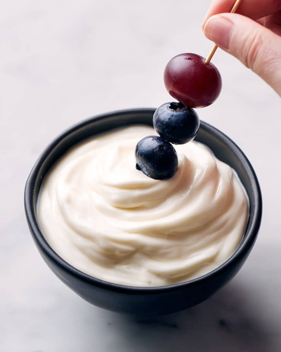 A black bowl filled with a thick, creamy white yogurt-like layer that is smoothly spread with gentle swirls. On top, a skewer held by a woman's hand is placed vertically into the cream, with one dark red grape at the top and three shiny dark blue blueberries stacked beneath it. The bowl sits on a white marbled surface. photo taken with an iphone --ar 4:5 --v 7