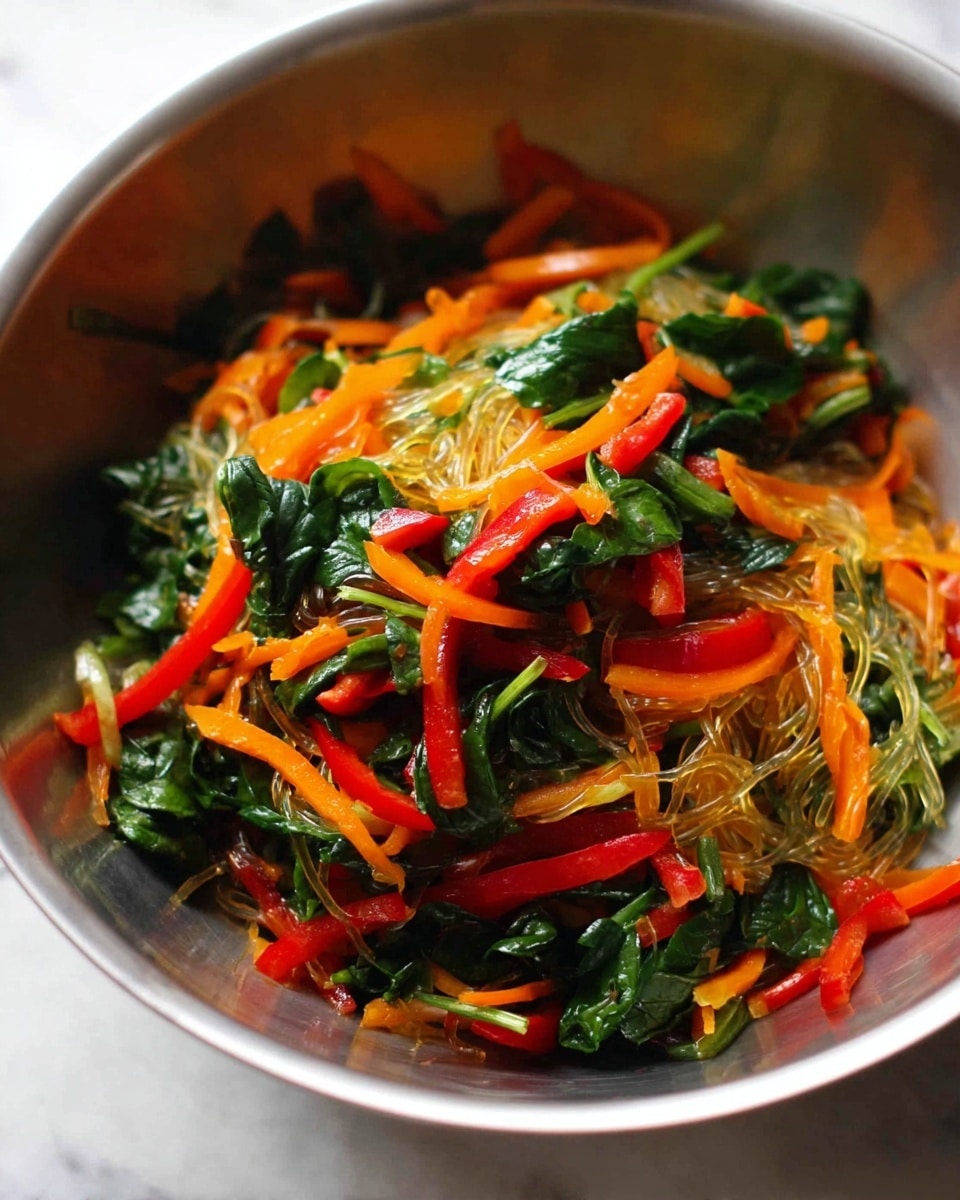 The image shows a metal bowl filled with a colorful vegetable mix. The base layer consists of a translucent tangle of glass noodles. On top of that, there is a mix of vibrant red strips of bell pepper, thin orange strips of carrot, and bright green spinach leaves, giving the dish a fresh and healthy look. The vegetables have a slightly wilted texture, suggesting they might be lightly cooked or sautéed. The bowl is placed on a white marbled surface. photo taken with an iphone --ar 4:5 --v 7