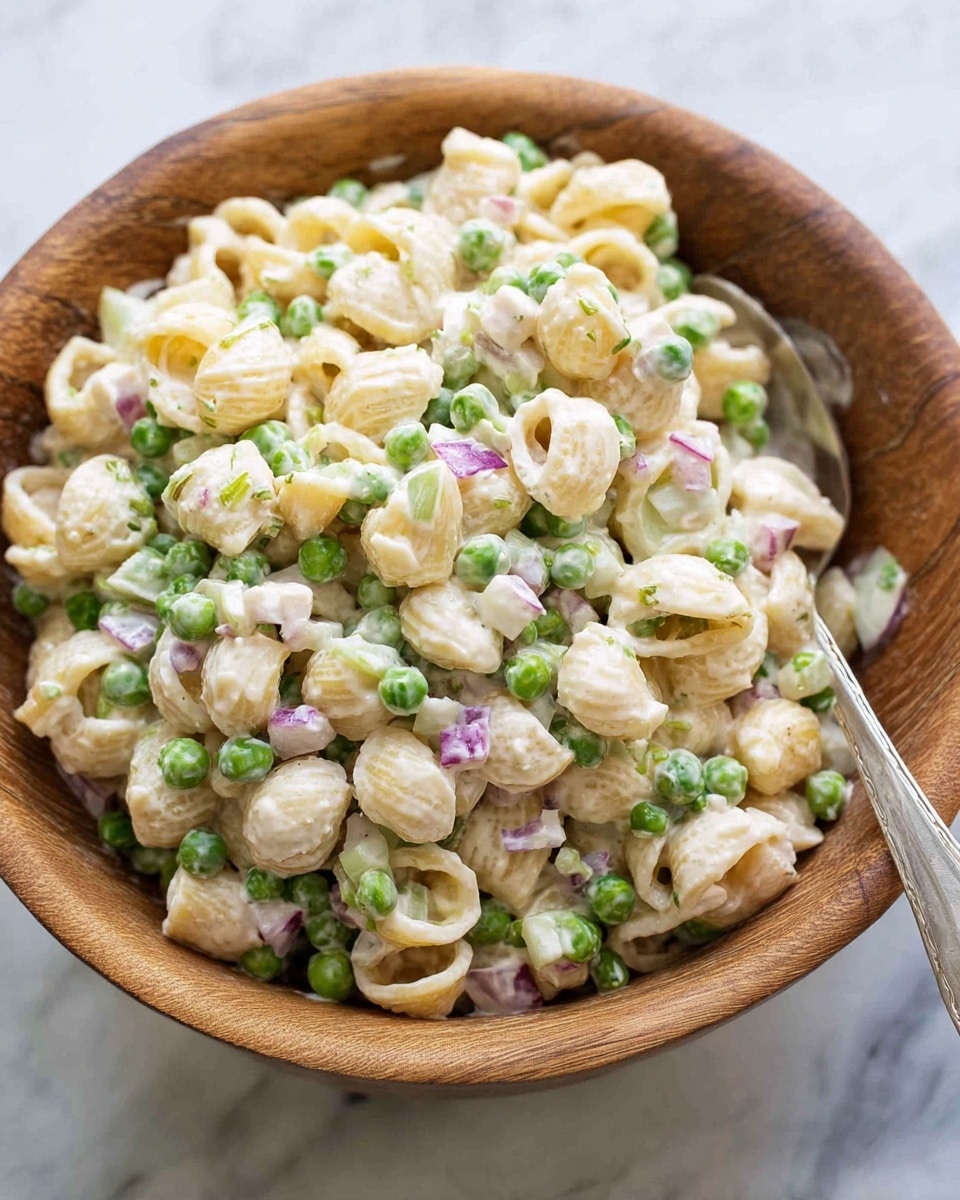 A large wooden bowl filled with creamy pasta salad sits on a white marbled surface. The salad has three main layers: small shell pasta coated with a smooth white dressing, bright green peas scattered evenly throughout, and pieces of light purple onion and pale green celery mixed in. The pasta looks soft and slightly shiny from the dressing, while the peas add a fresh pop of color. A metal spoon is placed inside the bowl, ready to serve. Photo taken with an iphone --ar 4:5 --v 7