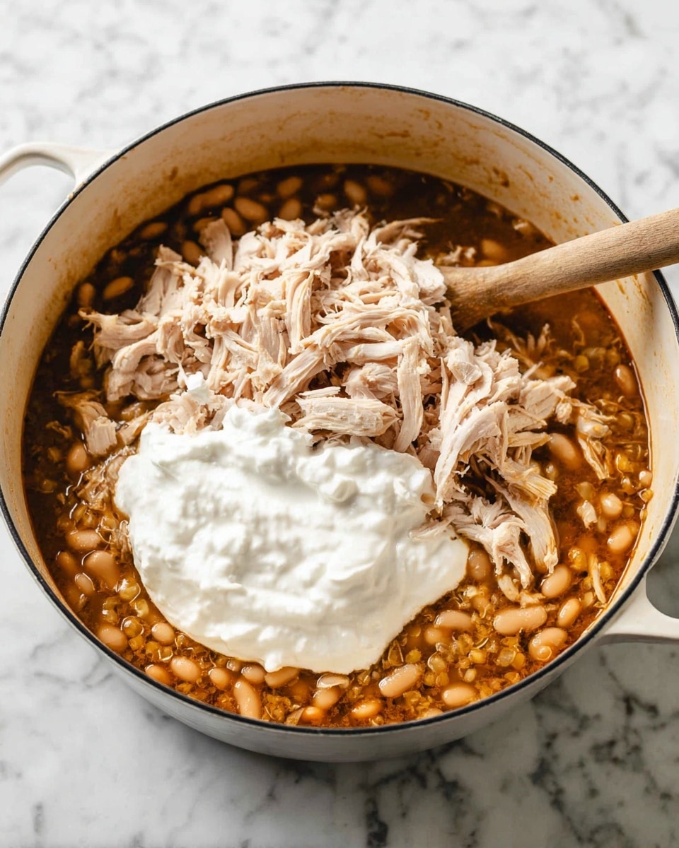 A white pot on a white marbled surface holds a dish with three main layers. The bottom layer is a brown sauce with a mix of light tan beans and grains. On top of that is a layer of shredded light pinkish turkey meat. The top layer is a dollop of thick white cream placed in the center. A wooden spoon rests inside the pot, reaching into the dish. Photo taken with an iphone --ar 4:5 --v 7