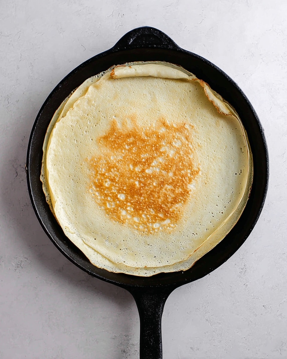 A single thin crepe lies flat in a black cast iron pan with a handle extending towards the bottom right. The crepe has a light creamy color with a center area showing a patch of golden brown where it is cooked more. The edges are slightly crisp and delicate, curling up a bit, revealing the thin and smooth texture. The pan rests on a white marbled surface, creating a clean and simple background. Photo taken with an iphone --ar 4:5 --v 7