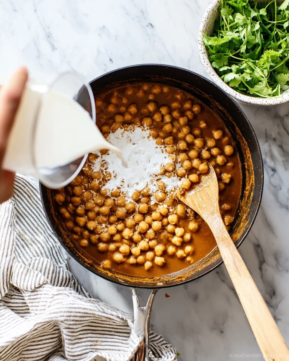 A black pot filled with a rich brown sauce at the bottom, layered with light beige chickpeas on top. White liquid, likely coconut milk, is being poured into the pot from the left by a woman's hand, creating a creamy white layer mixing slightly with the sauce. A wooden spoon with a light brown handle is resting inside the pot on the right side, partially covered in the brown sauce. The pot sits on a white marbled surface with a striped cloth underneath on the left and a white speckled bowl filled with fresh green leafy herbs at the top right. photo taken with an iphone --ar 4:5 --v 7