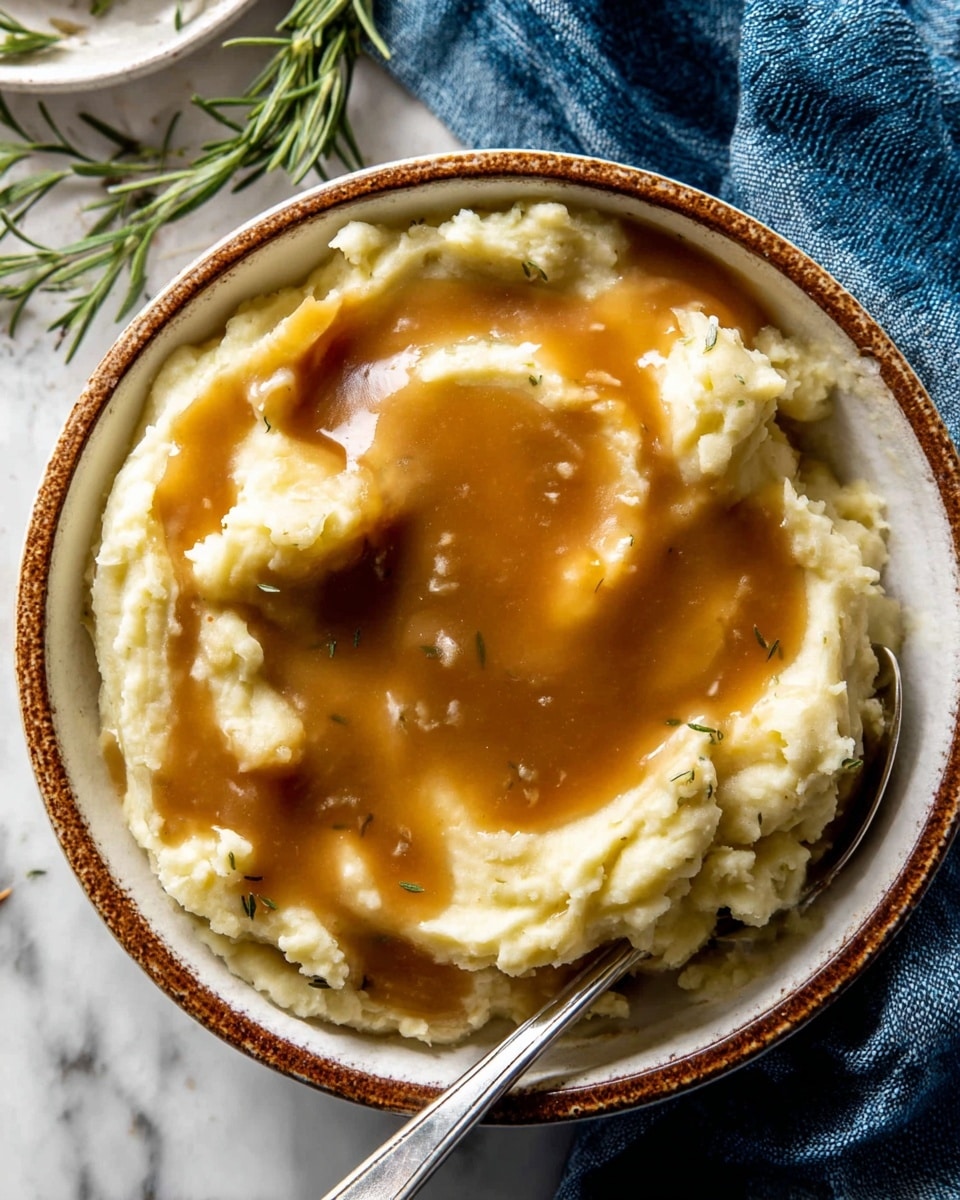 A close-up of a bowl filled with creamy mashed potatoes topped with a smooth, glossy brown gravy layer. The mashed potatoes form the bottom layer with a soft, slightly chunky texture in a pale yellow color. The gravy sits on top, spreading unevenly in a rich caramel brown shade with a shiny surface. The bowl is white with a rustic brown rim and a metal spoon rests inside on the right side. The setting includes a white marbled texture surface with a blue cloth and green rosemary sprigs visible near the top edge. Photo taken with an iphone --ar 4:5 --v 7