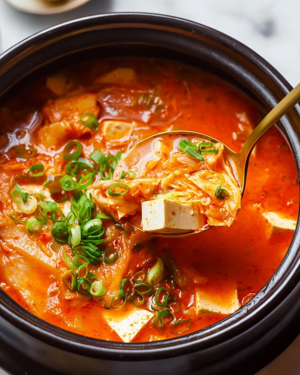 A black pot filled with a bright red-orange soup, showing several layers: at the bottom is a mix of kimchi and broth with a slightly thick texture, above that are chunky white tofu pieces with a soft, smooth texture, and scattered green sliced scallions on top adding color contrast. A gold spoon lifts a portion near the center, holding tofu, kimchi, and broth, emphasizing the rich, warm look of the soup. The pot is set on a white marbled surface. Photo taken with an iphone --ar 4:5 --v 7