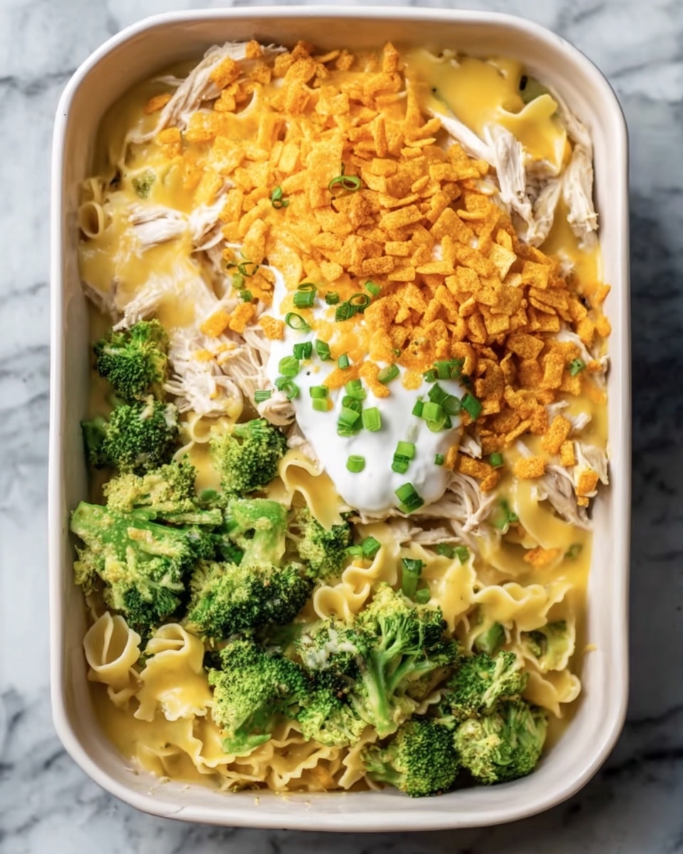 The dish is served in a beige rectangular baking dish placed on a white marbled surface. Inside, the bottom layer consists of broad flat noodles, partially covered by bright green broccoli florets scattered mainly around the edges. On one side, there is a generous serving of shredded light brown chicken arranged loosely. The top is mostly covered with a smooth, pale yellow cheese sauce. Bright orange shredded cheese sprinkles over half of the dish, atop which small chopped green herbs add a fresh contrast. In the middle, a dollop of white sour cream sits prominently. photo taken with an iphone --ar 4:5 --v 7