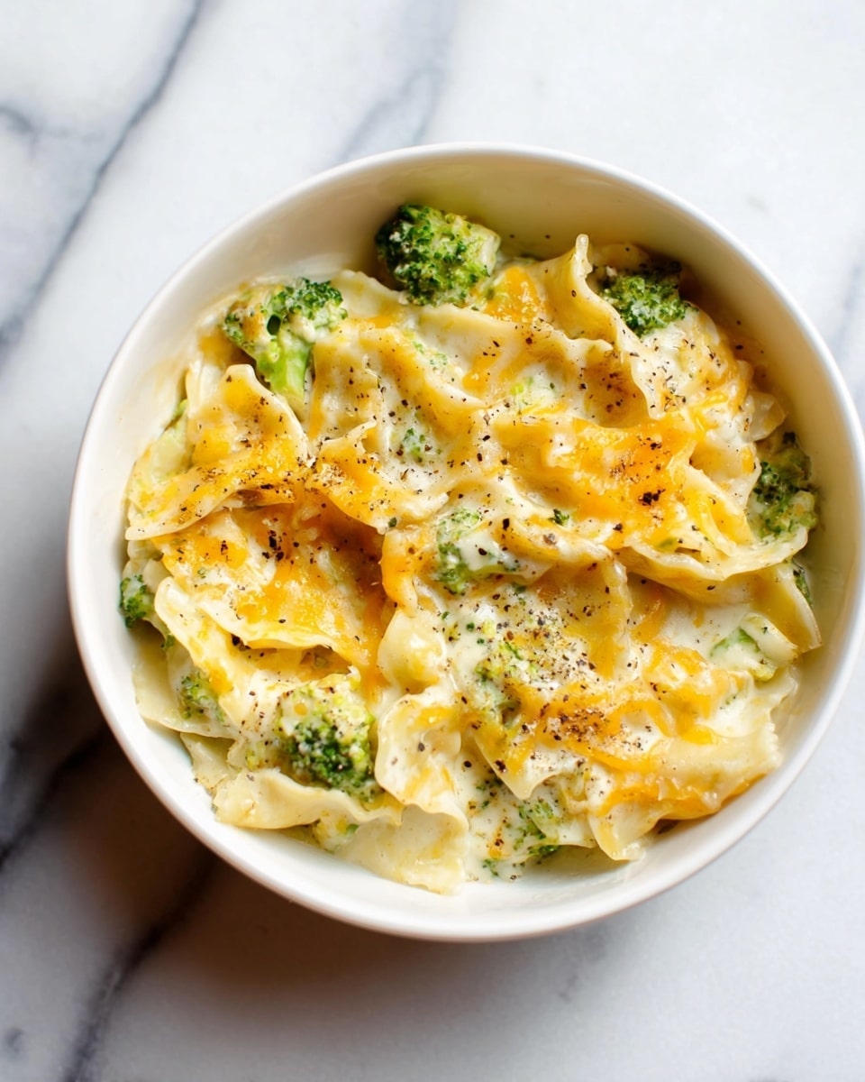 A white bowl filled with a creamy pasta dish featuring wide, folded noodles mixed with small pieces of bright green broccoli. The pasta is generously coated in a smooth, light yellow cheese sauce with melted orange cheese patches on top, sprinkled with small black pepper specks. The bowl sits on a white marbled surface, showing a cozy, warm meal ready to be served. photo taken with an iphone --ar 4:5 --v 7