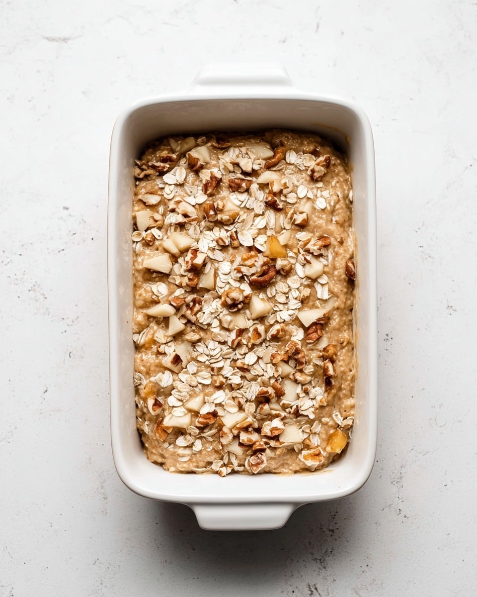 A white rectangular baking dish filled with a thick, lumpy batter that has a light brown color. The batter is mixed with pieces of chopped nuts and fruits, visible as off-white and light brown chunks scattered throughout. On top, there is a layer of raw oats and more whole nuts sprinkled unevenly, adding texture and contrast with their beige and tan colors. The dish sits on a white marbled surface. photo taken with an iphone --ar 4:5 --v 7