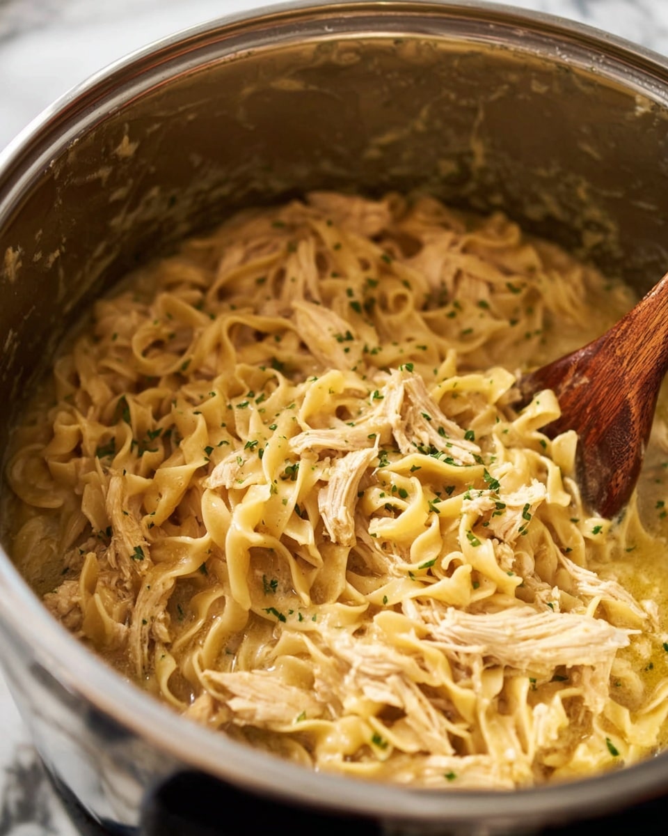 A close-up of a silver pot filled with creamy cooked egg noodles mixed with shredded light brown chicken pieces, all coated in a smooth yellowish sauce sprinkled with small bits of green herbs. A wooden spoon with a blurred handle is partially submerged, stirring the thick mixture inside the pot. The background shows a white marbled texture surface. photo taken with an iphone --ar 4:5 --v 7
