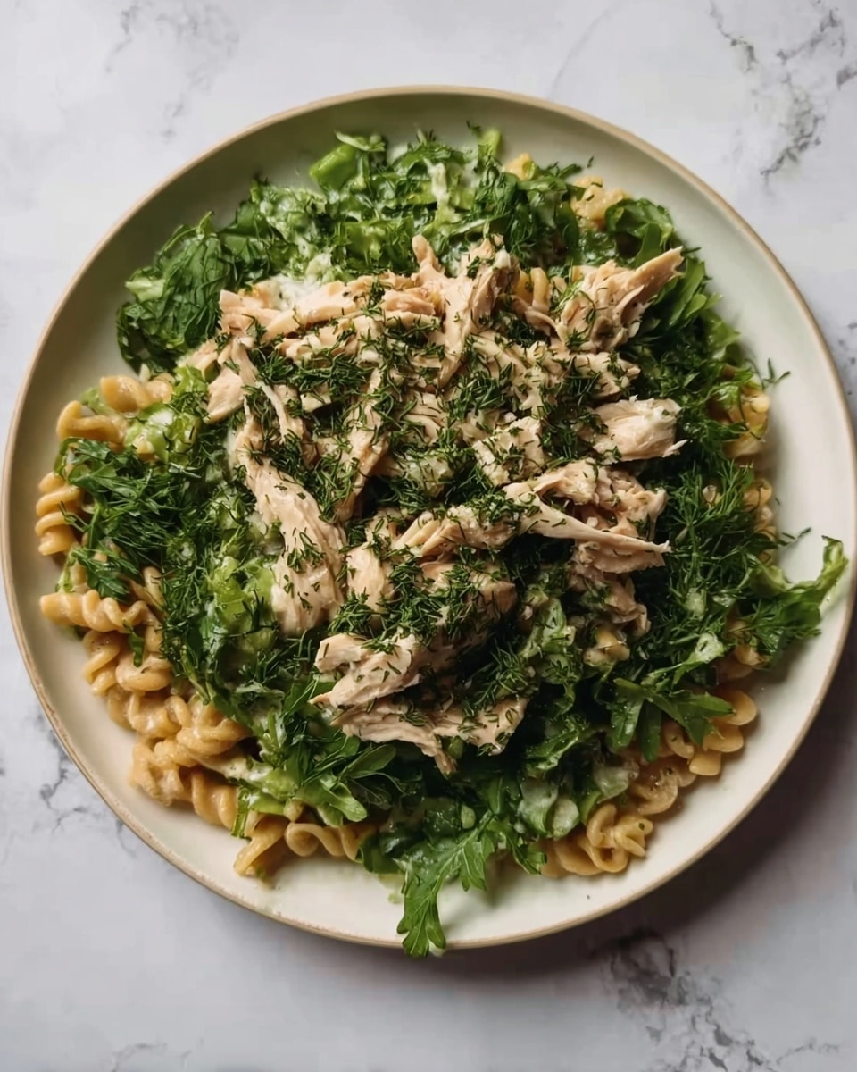 A white plate with pasta at the bottom layer, showing a light golden-brown color and slightly curly shape. The middle layer is fresh green leafy vegetables spread over the pasta, creating a lush green base. On top, there are pieces of shredded light beige chicken scattered evenly. The dish is garnished with finely chopped herbs, giving a touch of dark green color over the chicken and vegetables. The plate is placed on a white marbled surface. photo taken with an iphone --ar 4:5 --v 7