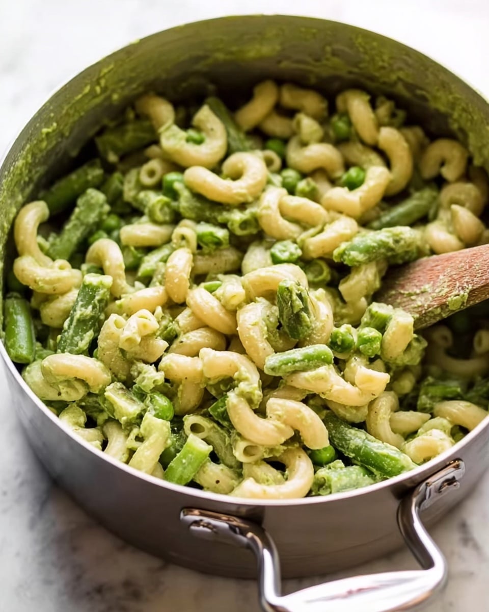 The image shows a pot filled with elbow-shaped pasta mixed with green vegetables like peas and green beans, all coated in a light green sauce that looks creamy. The pasta is curved and looks soft, with some pieces sticking together with the sauce. A wooden spoon is partially visible on the right, stirring the mixture inside the pot. The pot is silver with a shiny handle, and the scene is set on a white marbled surface. The colors mainly include pale yellow from the pasta and different shades of green from the sauce and vegetables. photo taken with an iphone --ar 4:5 --v 7