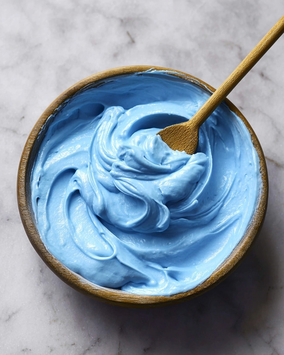 A round wooden bowl filled with a creamy, smooth blue mixture with swirls and soft peaks showing its thick texture. A wooden spoon is dipped in the mixture, resting in the bowl with some of the blue cream lifted slightly. The bowl is placed on a white marbled surface, adding contrast to the vivid blue mixture inside. The lighting highlights the smoothness and thickness of the blue cream, making it look rich and soft. Photo taken with an iphone --ar 4:5 --v 7