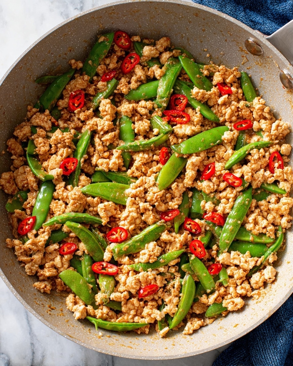 The image shows a light gray pan filled with cooked crumbled turkey that is light brown and has a soft, crumbly texture. Scattered among the turkey are bright green snap peas, some halved, which add a fresh and crisp look. Thin slices of red chili pepper pieces are spread evenly, adding small spots of vibrant red throughout the pan. The pan sits on a white marbled surface with a bit of blue fabric partially visible nearby. The mix of colors gives a fresh and appetizing feel to the dish photo taken with an iphone --ar 4:5 --v 7