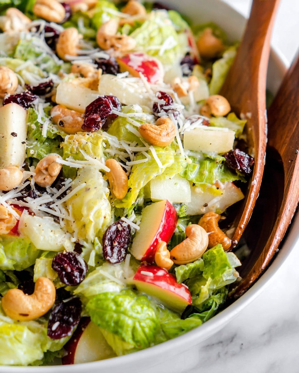 A close-up view of a fresh salad in a white bowl on a white marbled surface, showing layers starting with green crisp lettuce leaves forming the base, topped with light brown cashew nuts, small chunks of red and yellow apple pieces, and white shredded cheese scattered all over. There are also dark red dried cranberries mixed evenly throughout. A wooden salad spoon is partially visible on the right side, scooping the salad. The salad is lightly dressed with a creamy dressing that glistens on the ingredients. Photo taken with an iphone --ar 4:5 --v 7