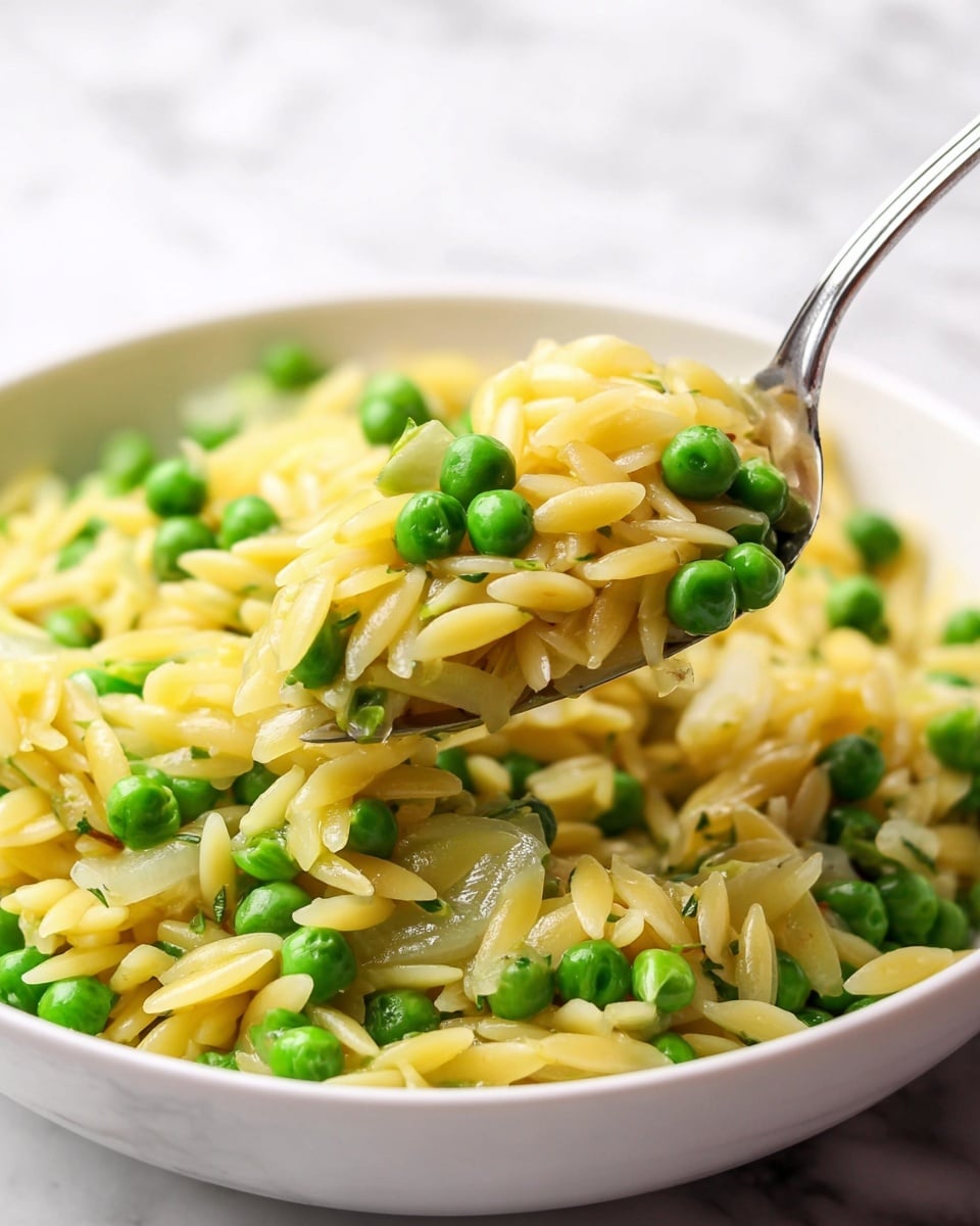 The image shows a close-up of a white bowl filled with a dish that has three main layers mixed together: small yellow orzo pasta, bright green peas, and translucent pieces of cooked onion. The orzo pasta looks soft and shiny, evenly mixed with the round green peas that add a pop of color. The translucent onion pieces are scattered throughout, adding texture. A silver fork is lifting a portion of the dish, with the spoon holding a balanced mix of orzo, peas, and onion. The bowl is set on a white marbled surface, highlighting the colors and texture of the food. Photo taken with an iphone --ar 4:5 --v 7