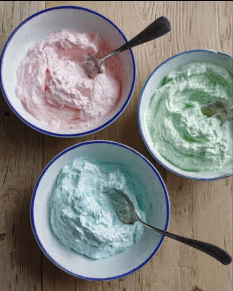 Three white bowls with blue rims sit on a wood surface. Each bowl holds a different colored soft creamy substance with a smooth, fluffy texture. The top bowl has a light pink cream, the bottom left bowl has a pale green cream, and the bottom right bowl has a light blue cream. Each bowl contains a shiny silver spoon resting inside the cream. The scene has a calm and simple look with soft colors. photo taken with an iphone --ar 4:5 --v 7