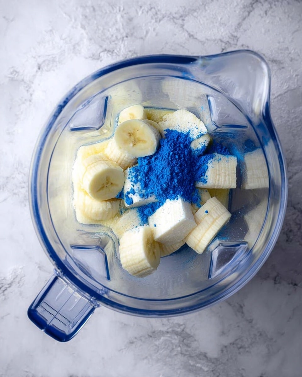 The image shows a clear blender jar placed on a white marbled surface. Inside the blender, there are several frozen banana pieces that are pale yellow and slightly frosty, with some creamy white cubes that have a firm texture. On top of these ingredients, there is a small pile of bright blue powder with a fine texture, slightly scattered around. The blender jar has a handle on the side, and the whole scene is brightly lit, giving a clean and fresh look. photo taken with an iphone --ar 4:5 --v 7
