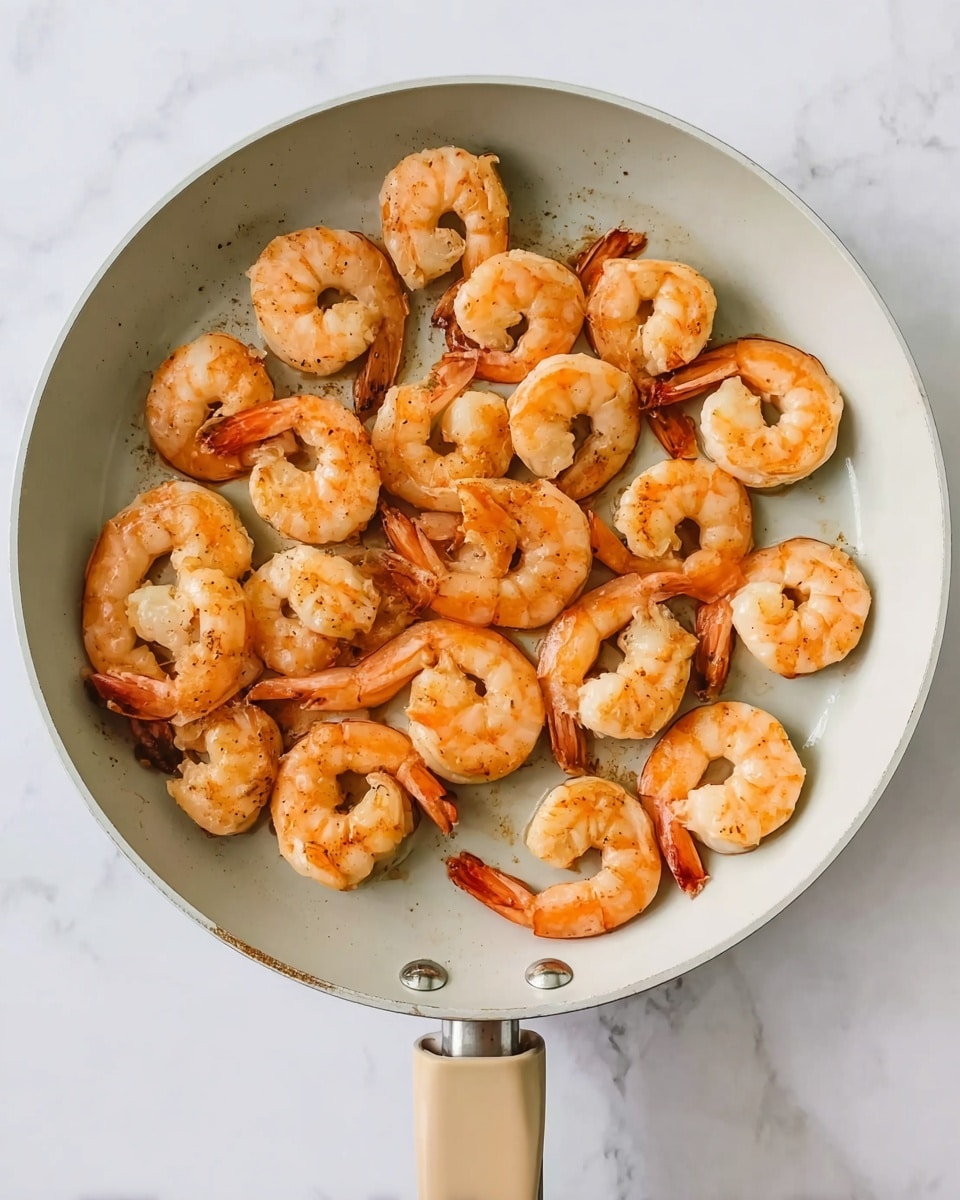 The image shows a round, white frying pan filled with about 30 cooked shrimp arranged in a single layer, each shrimp curved and displaying a light orange-pink color with some slight sear marks. The shrimp have a smooth, slightly shiny texture, and their tails are still attached. The frying pan is held by a beige handle and sits on a white marbled surface. photo taken with an iphone --ar 4:5 --v 7