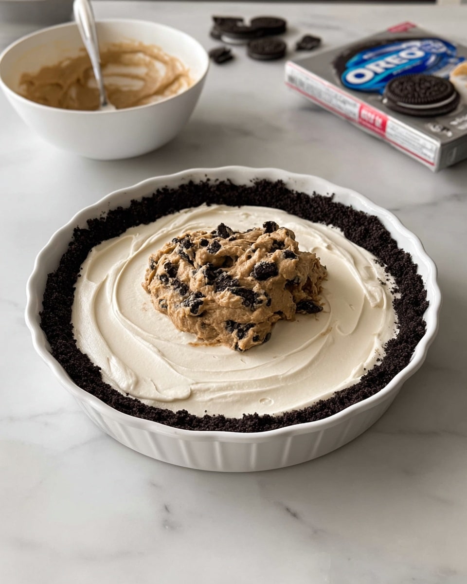 A white pie dish holds a dark brown, crumbly Oreo cookie crust forming the bottom and sides. Inside the crust is a smooth, thick and creamy white layer spread evenly across the base. On top of that sits a mound of light brown dough-like mixture with small dark chunks mixed in, placed just off-center. The pie dish rests on a white marbled surface. In the background, there is a white bowl with some leftover light brown mixture and a spoon inside it, along with an opened pack of Oreo cookies. photo taken with an iphone --ar 4:5 --v 7