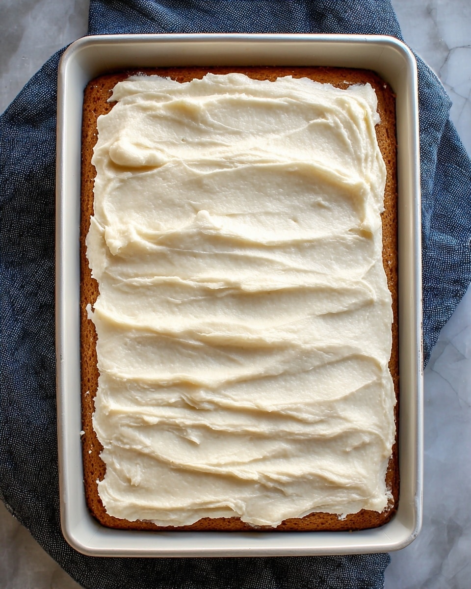 The image shows a rectangular cake in a white rectangular pan, with two main layers visible. The bottom layer is a light brown cake with a soft, spongy texture. The top layer is a thick, evenly spread creamy white frosting that has smooth, slightly wavy lines and a soft, fluffy look. The pan sits on a dark blue cloth over a white marbled surface. The overall look is neat and simple, with the cake filling almost the entire pan. photo taken with an iphone --ar 4:5 --v 7