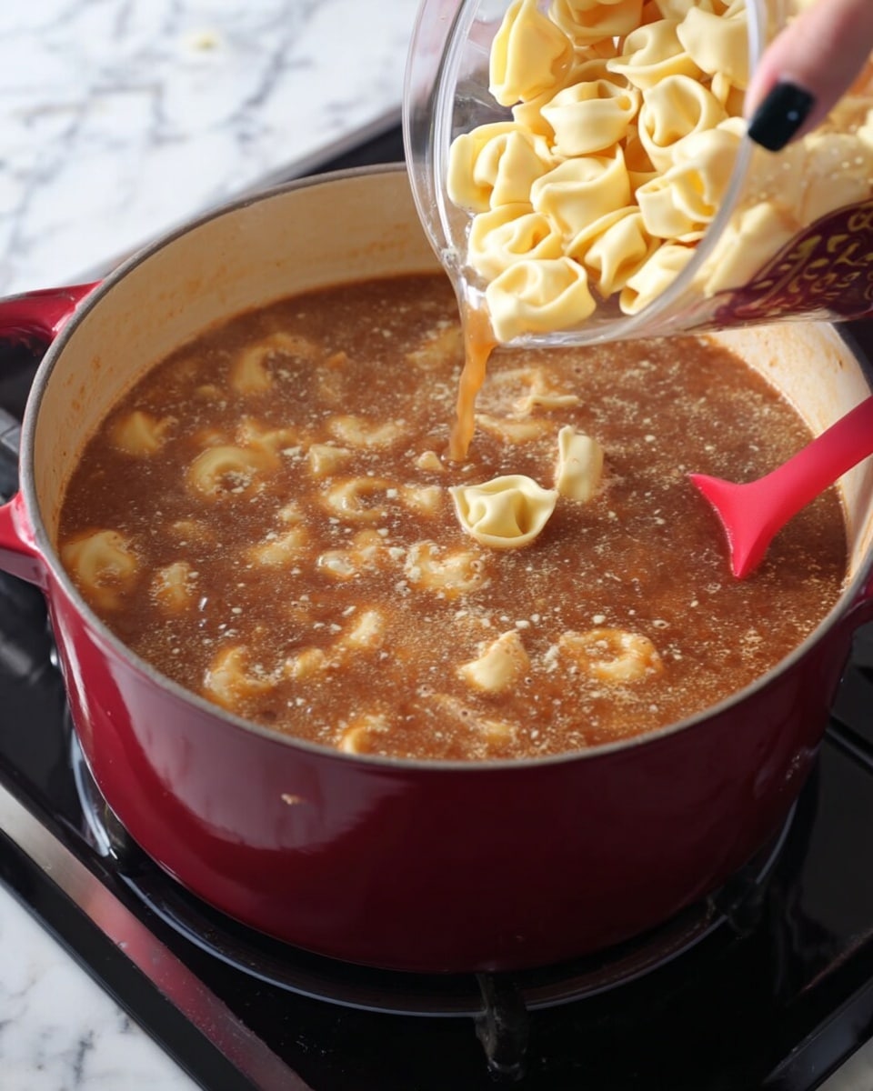 A deep red pot filled with a brown, bubbling soup sits on a black stovetop with a red spatula resting inside. Above the pot, a woman's hand with black painted nails is pouring pale yellow, uncooked tortellini pasta into the soup. The soup has a slightly frothy and thick texture with visible small bits of ingredients, and the pasta pieces contrast softly against the rich broth. The background is a white marbled texture. photo taken with an iphone --ar 4:5 --v 7
