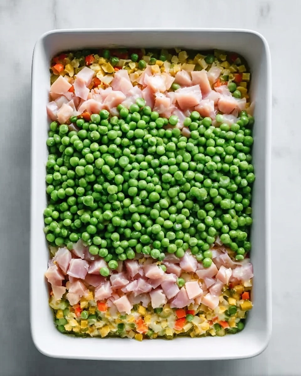 The dish is shown in a white rectangular baking dish on a white marbled surface. The bottom layer has small pieces of yellow, orange, and green vegetables mixed together. Above that is a layer of small, light pink pieces arranged evenly. The top layer is fully covered with bright green peas scattered all over. The dish looks fresh and colorful. Photo taken with an iphone --ar 4:5 --v 7