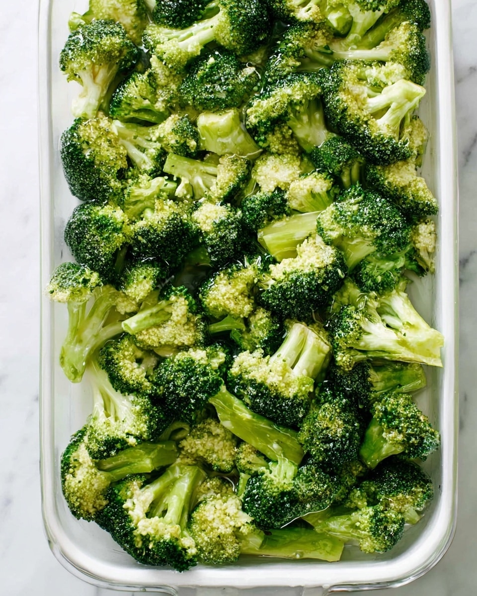 A large white rectangular dish filled with a single layer of fresh broccoli florets. The broccoli is bright green with a firm texture, showing the floret tops and thick stems evenly spread out. Some water or light dressing glistens on the broccoli pieces, and the dish rests on a white marbled surface. Photo taken with an iphone --ar 4:5 --v 7