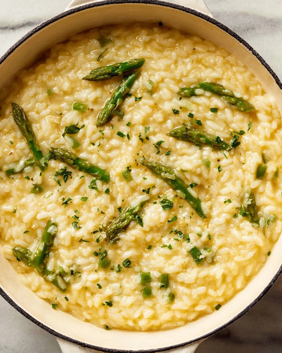 The dish is creamy risotto served in a white pot with a hint of black around the rim, placed on a white marbled surface. The risotto has soft yellow rice with a smooth, creamy texture that is mixed evenly throughout. Scattered inside the risotto are small, bright green asparagus pieces that add a fresh and slightly crisp look. Small bits of finely chopped green herbs are sprinkled on top, providing specks of deeper green color over the creamy yellow base. The risotto fills the pot almost to the top, showing some slight steam and shine from its moist texture. photo taken with an iphone --ar 4:5 --v 7