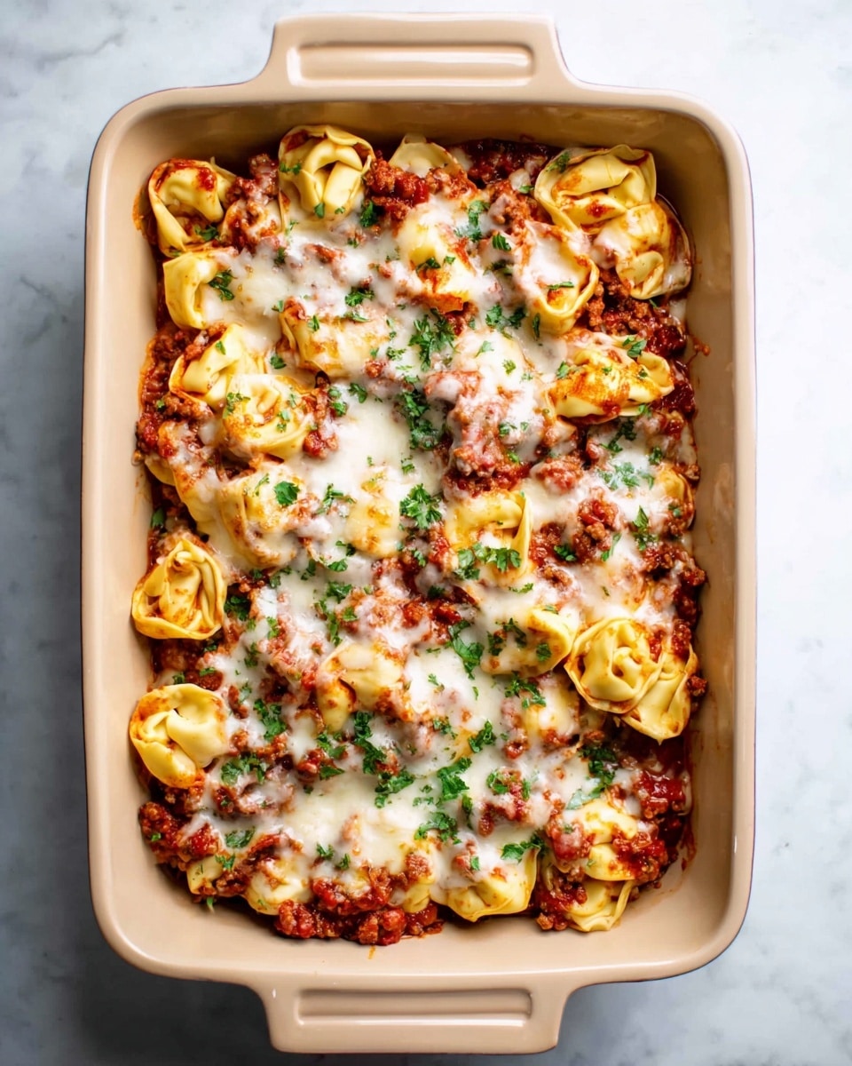 A beige rectangular baking dish filled with one layer of cooked tortellini pasta covered in red tomato meat sauce mixed with browned ground meat, topped with melted white cheese scattered evenly, and garnished with small green parsley leaves. The tortellini are folded in round shapes with visible sauce and cheese melting between them. The dish rests on a white marbled surface. photo taken with an iphone --ar 4:5 --v 7