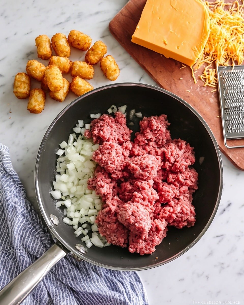 A black pan with a silver handle sits on a white marbled surface with a blue and white striped cloth nearby. Inside the pan are two main layers: the bottom layer is small white onion pieces, and the top layer is raw ground meat with a pink and red color, grouped in soft clumps. On the white marbled surface near the top right corner, a wooden board holds a block of orange cheese with some of it shredded beside a metal grater. At the top left on the marble surface, a small pile of golden-brown tater tots is arranged. A wooden spatula handle is partially visible on the left side. Photo taken with an iphone --ar 4:5 --v 7