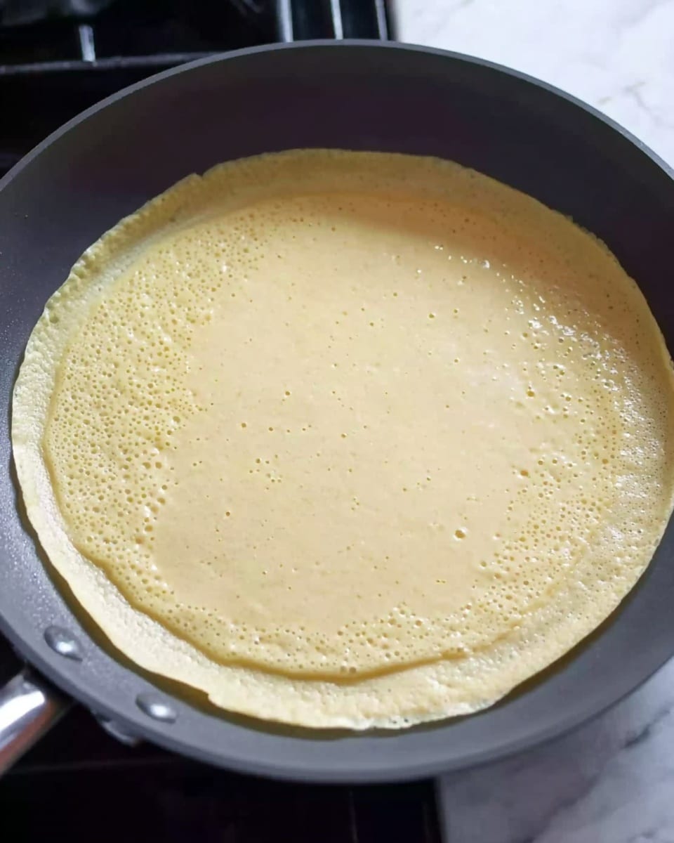 A thin, round crepe is cooking in a dark gray non-stick frying pan. The crepe is pale yellow with a smooth texture and small bubbles forming on the surface. The crepe edges are slightly lifted from the pan, indicating it’s cooking gently. The pan is placed on a stove with black grates visible around it. The scene shows just one layer: the crepe batter spreading evenly across the pan. The background includes a white marbled surface where the stove rests. Photo taken with an iphone --ar 4:5 --v 7