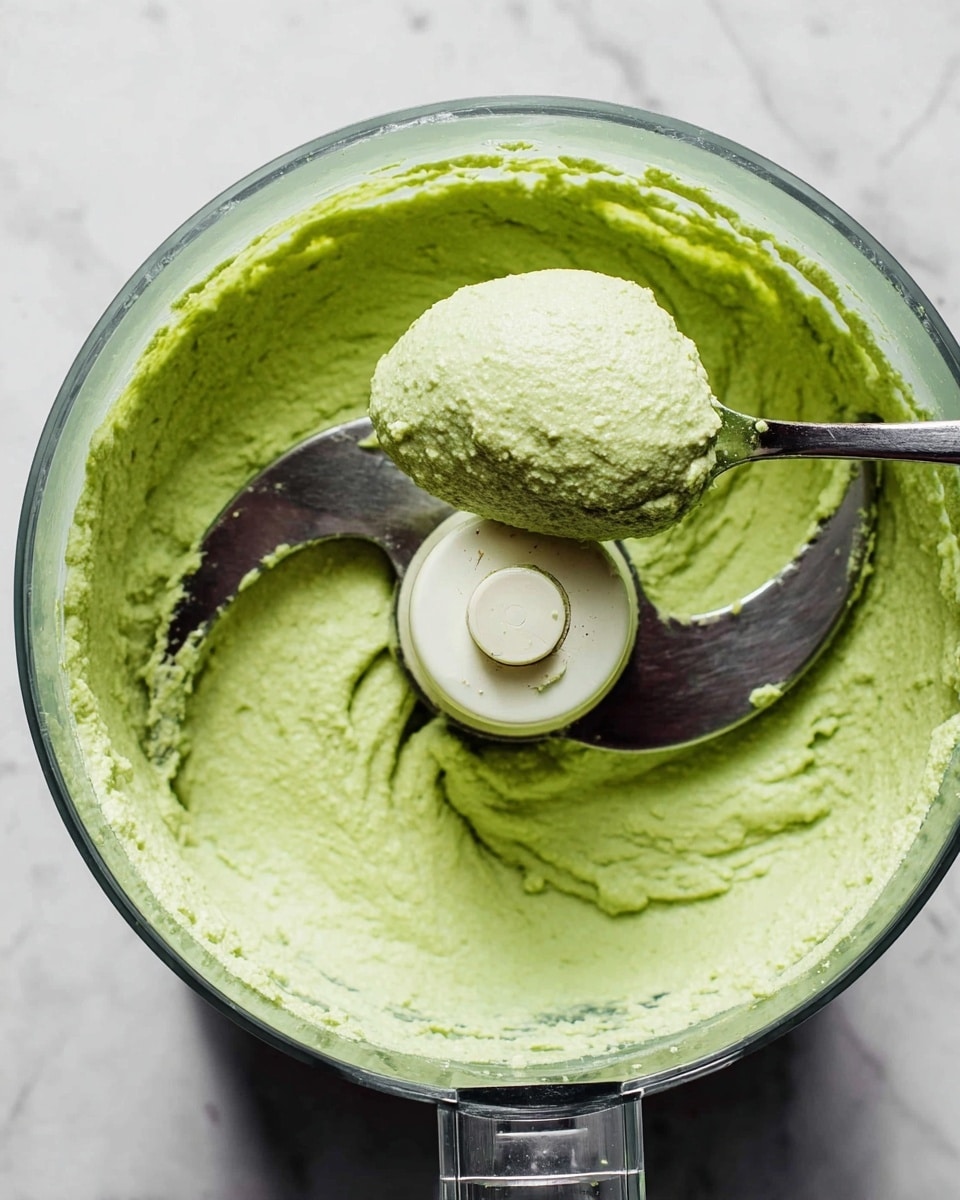 A clear food processor bowl filled with a smooth, thick, pale green paste with some light texture. A metal spoon inside the bowl holds a rounded scoop of the paste, showing its creamy and slightly lumpy consistency. The food processor blade is visible in the center, surrounded by the spread-out green mixture. The background is a white marbled texture. Photo taken with an iphone --ar 4:5 --v 7