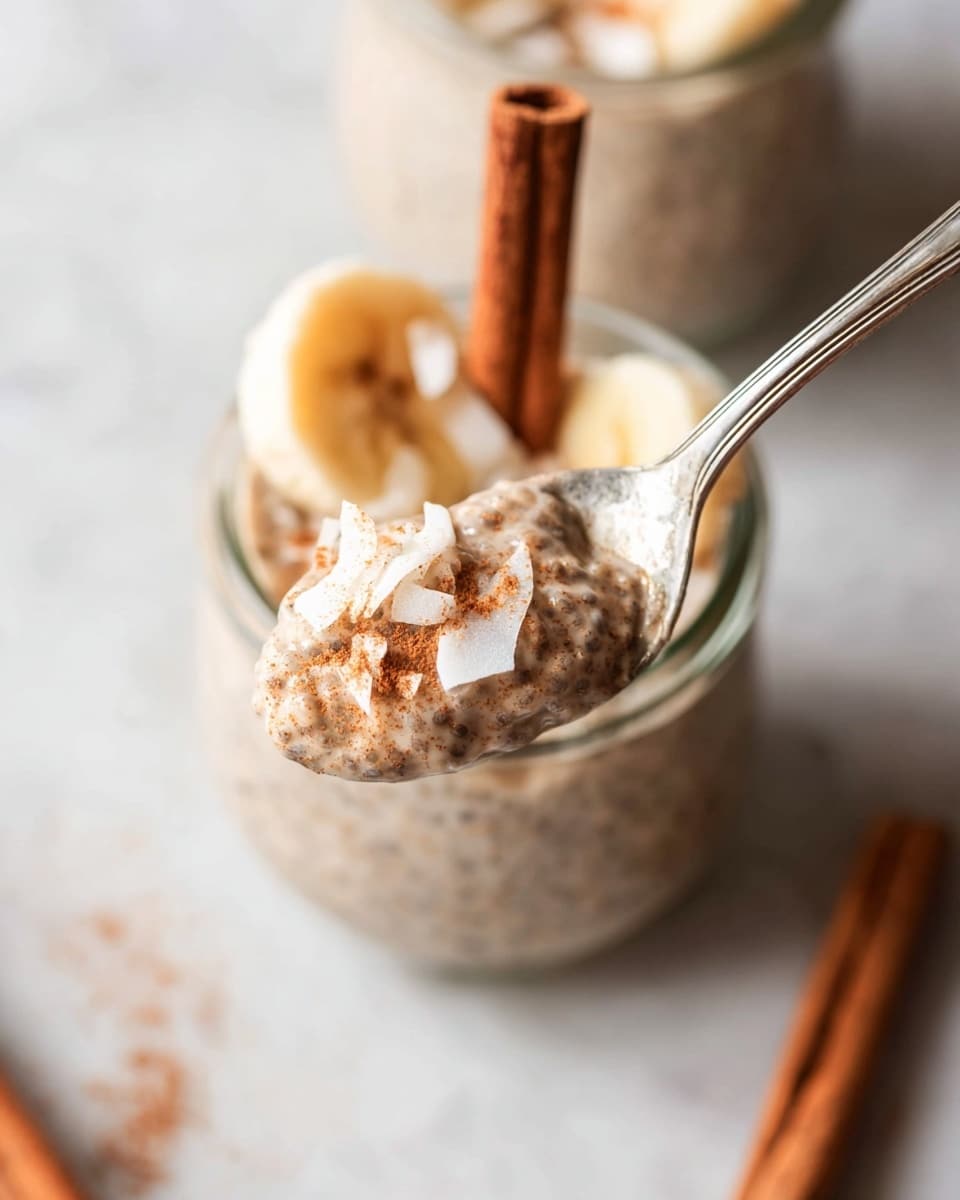 A close-up view of a spoon holding a thick creamy chia seed pudding that is light brown with visible chia seeds, topped with small white coconut flakes and a light dusting of cinnamon powder. In the background, a transparent glass jar shows layers of the same pudding with a round slice of light yellow banana on top, some extra coconut flakes, and a cinnamon stick standing upright in the pudding. The setting is on a white marbled surface, with another cinnamon stick lying flat out of focus. The photo taken with an iphone --ar 4:5 --v 7