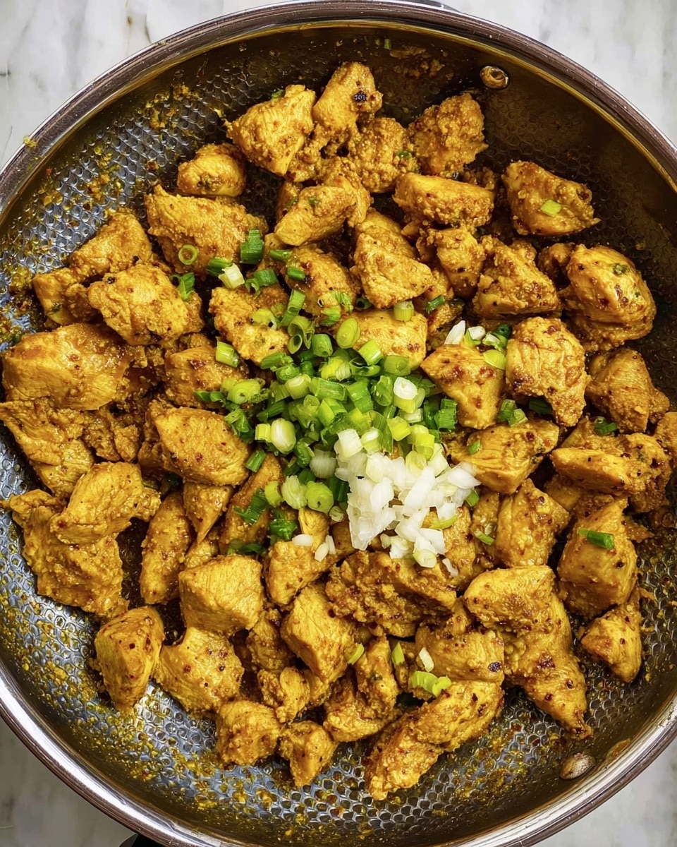 A frying pan with a silver edge holds many pieces of cooked chicken in a yellow-brown spice mix. The chicken pieces show a rough texture with small bits of cooked spices. On top near the center, there is a small pile of white minced garlic and bright green chopped green onions. The pan is sitting on a surface with a white marbled texture. The photo taken with an iphone --ar 4:5 --v 7