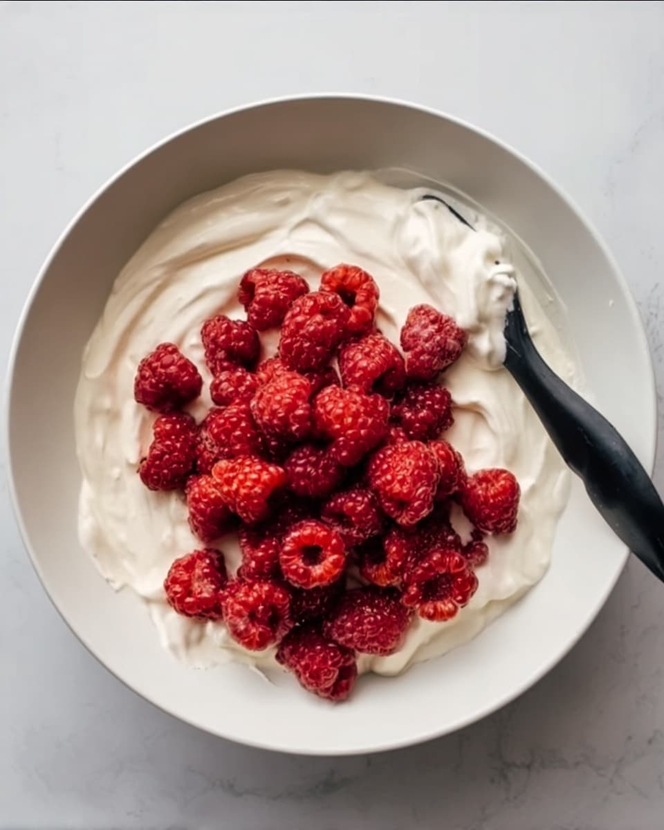 A white bowl filled with a smooth, thick white cream spread evenly on the bottom with a pile of fresh, bright red raspberries placed in the center. A black spatula rests on the right side of the bowl, partially covered with cream. The bowl sits on a white marbled surface. Photo taken with an iphone --ar 4:5 --v 7