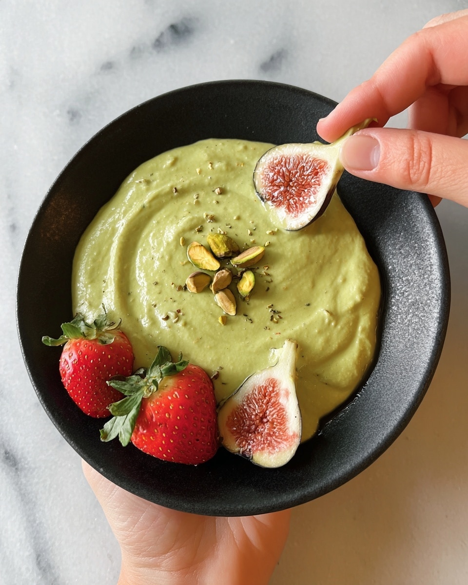 A black bowl holds a smooth, creamy green dip spread evenly in the center with a few whole pistachios on top. Along the edge of the bowl, there are fresh strawberries and halved figs with their pinkish interiors facing up. A woman's hand is holding one fig half dipped partway into the green dip. The bowl is set on a white marbled surface. photo taken with an iphone --ar 4:5 --v 7
