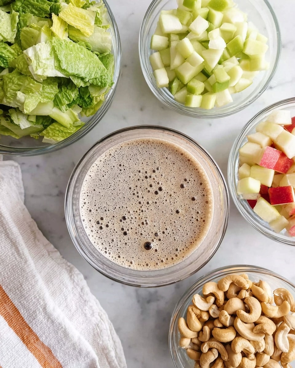 The image shows five clear glass bowls arranged on a white marbled surface. The central bowl contains a frothy, light brown liquid with bubbles on top. The top left bowl is filled with fresh, bright green romaine lettuce pieces. The top right bowl holds small, diced pieces of green pear. The bottom right bowl has a mix of small cubed red apple pieces. The bottom center bowl contains a pile of whole cashew nuts, light brown in color with smooth textures. A white cloth with thin orange stripes is placed on the left edge. Photo taken with an iphone --ar 4:5 --v 7