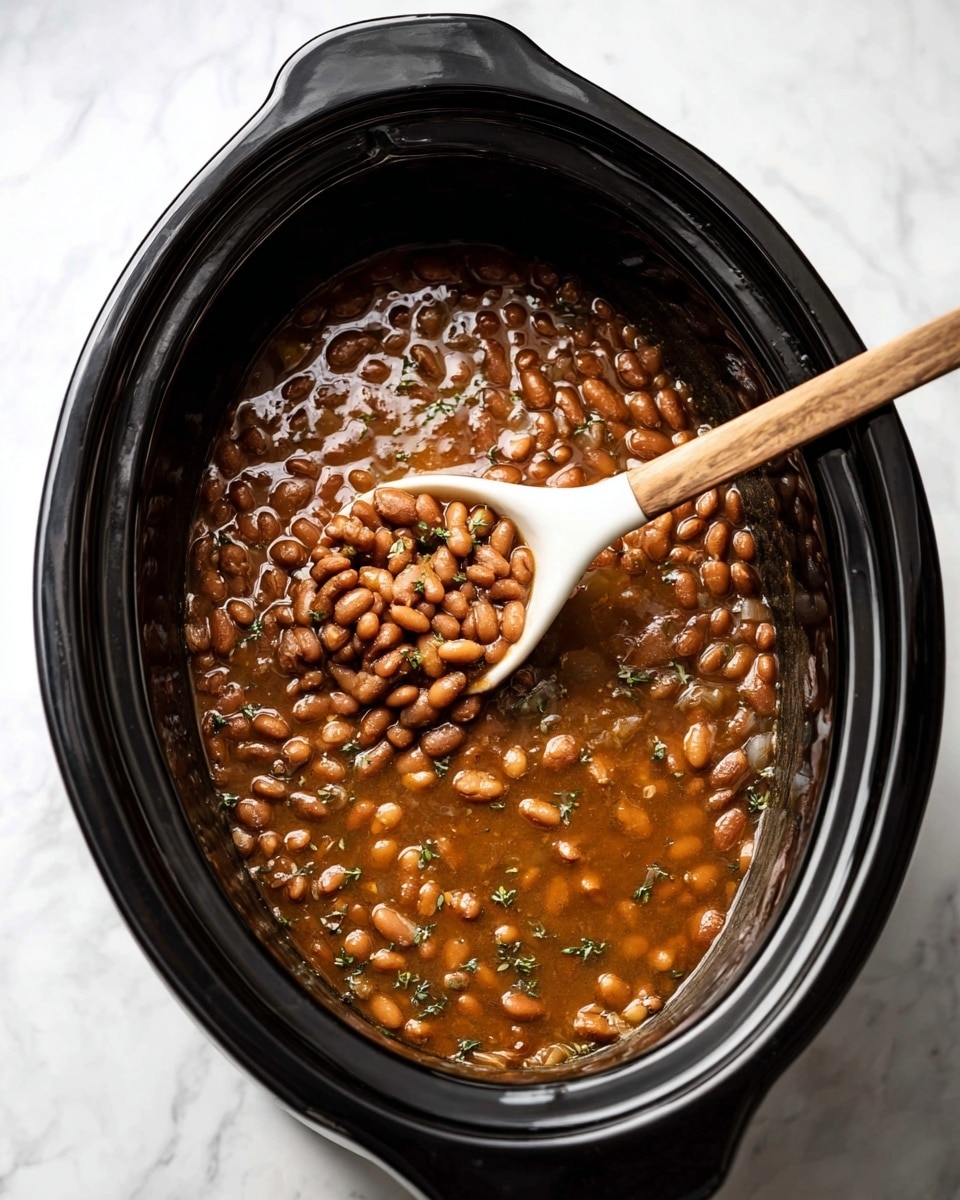 A black oval slow cooker is filled with cooked brown beans in a thick, shiny light brown sauce with small pieces of green herbs and onion mixed in. A white spoon with a light wooden handle is stirring the beans, showing some beans lifted and resting on the spoon's bowl. The slow cooker is placed on a white marbled surface, and the photo is taken from above. photo taken with an iphone --ar 4:5 --v 7