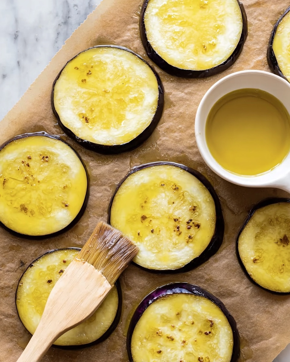 There are nine round, yellowish eggplant slices with visible seeds and some oil spread on the top surfaces, arranged on a brown parchment paper. A small white cup filled with golden oil is placed near the top right corner. A brush with a light wooden handle and dark bristles is touching one eggplant slice near the bottom left. The eggplant skin is dark purple, thinly bordering each slice. The background is a white marbled texture. Photo taken with an iphone --ar 4:5 --v 7