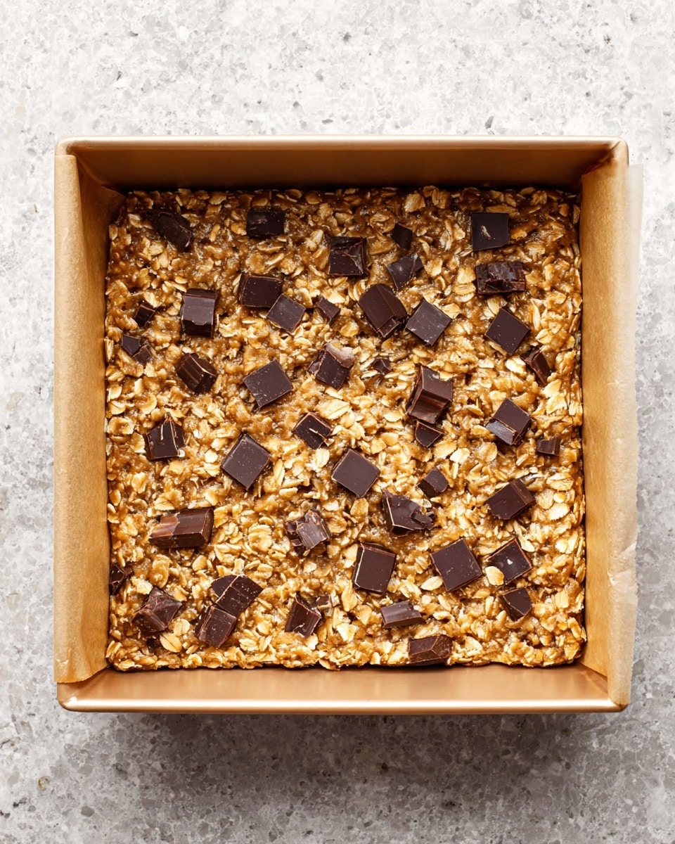 The image shows a single layer of oatmeal cookie dough pressed evenly inside a square baking pan with parchment paper lining the sides. The dough is golden brown with a rough texture formed by rolled oats mixed throughout, and scattered dark brown chunks of chocolate evenly spread on top. The pan sits on a white marbled surface. Photo taken with an iphone --ar 4:5 --v 7