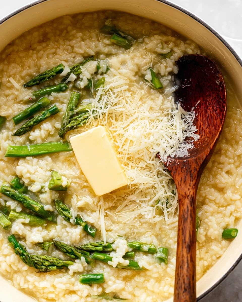 A close-up view of creamy risotto in a white pot, showing three clear layers: the creamy rice as the base in off-white with visible grains, bright green asparagus tips mixed throughout the rice, and a smooth, light yellow square of melting butter placed near the center, topped with a small pile of finely grated pale yellow cheese. A dark wooden spoon with a shiny, slightly wet surface rests on the right side in the risotto. The pot sits on a white marbled surface. Photo taken with an iphone --ar 4:5 --v 7