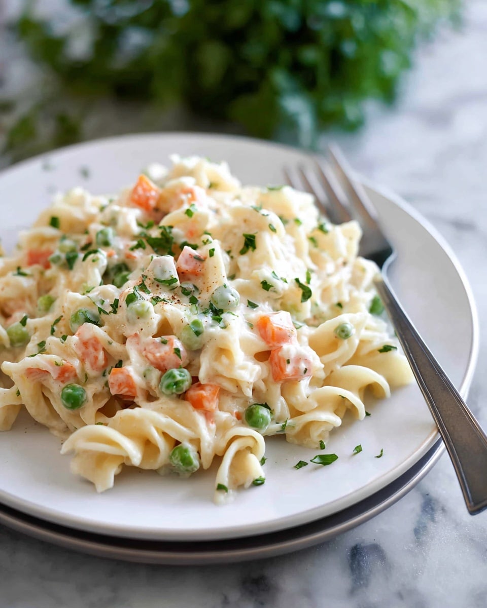 A white plate holds a creamy pasta dish made of three layers. The base layer shows curly, pale beige egg noodles with a soft texture. On top of the noodles is a thick layer of creamy white sauce mixed with small cubes of orange carrots and bright green peas. Scattered across the dish are small bits of green herbs adding a fresh touch. A silver fork rests on the right side of the plate, and the white plate sits on a white marbled surface with blurred green plants in the background. photo taken with an iphone --ar 4:5 --v 7