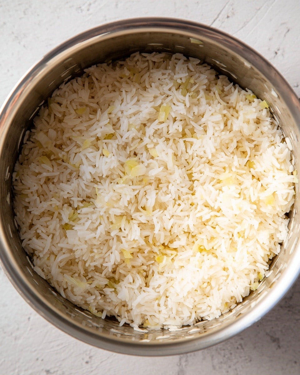 A close-up view of a stainless steel pot filled with uncooked white rice mixed with small pieces of light yellow chopped onions. The rice grains are long and separated, covering the bottom of the pot evenly, with a few onions scattered throughout. The pot's inner surface is shiny and reflective, showing the texture of the grains and onions clearly. The background surface is a white marbled texture. photo taken with an iphone --ar 4:5 --v 7