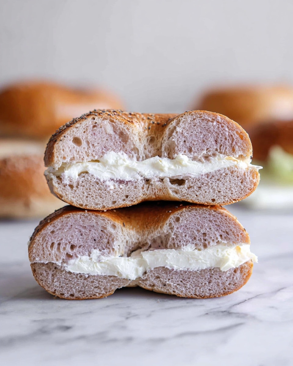 Two halves of a light purple bagel stacked on each other are shown, each cut side facing forward, revealing a soft textured inside with small air holes. Between the two halves, there is a layer of white cream cheese spread evenly. The bagel’s crust is slightly browned and smooth. The background is a white marbled surface with a blurred glimpse of more bagels in the distance. The photo taken with an iphone --ar 4:5 --v 7