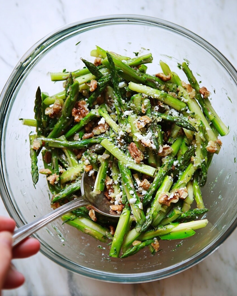 A clear glass bowl filled with green asparagus pieces that are thinly sliced into long strips and mixed with small brown walnut chunks, sprinkled with fine white cheese crumbles. A woman's hand holds a silver spoon resting on the edge of the bowl. The bowl is placed on a white marbled surface. photo taken with an iphone --ar 4:5 --v 7